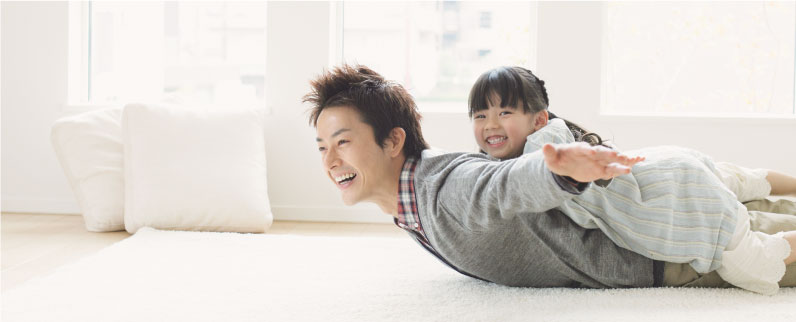写真：絨毯の上で両手を広げたお父さんの背中に女の子が乗って二人で遊んでいる